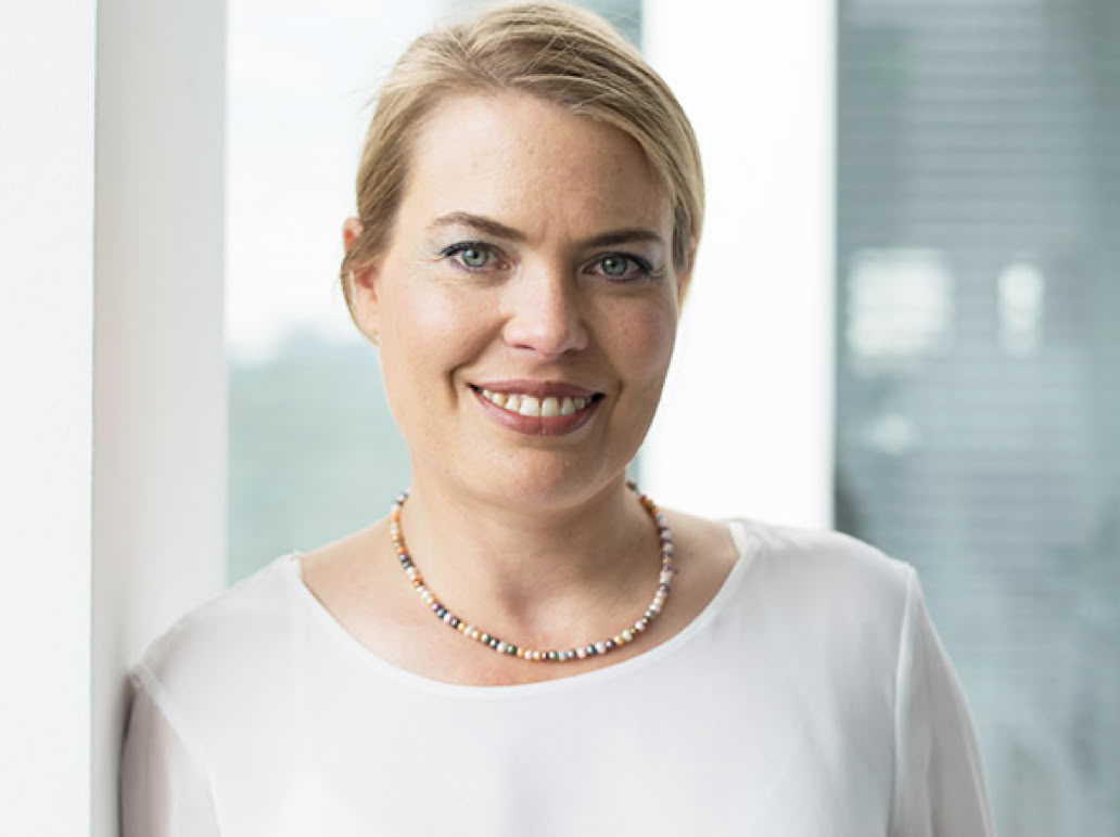 Eine lächelnde Frau mit blonden Haaren trägt eine weiße Bluse und eine Halskette, vor einem Fenster stehend.