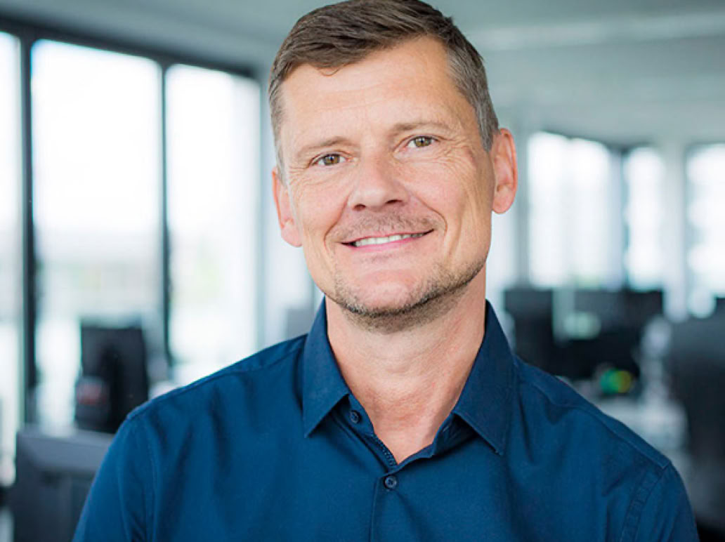 Ein lächelnder Mann in einem blauen Hemd steht in einem modernen Büro mit großen Fenstern im Hintergrund.