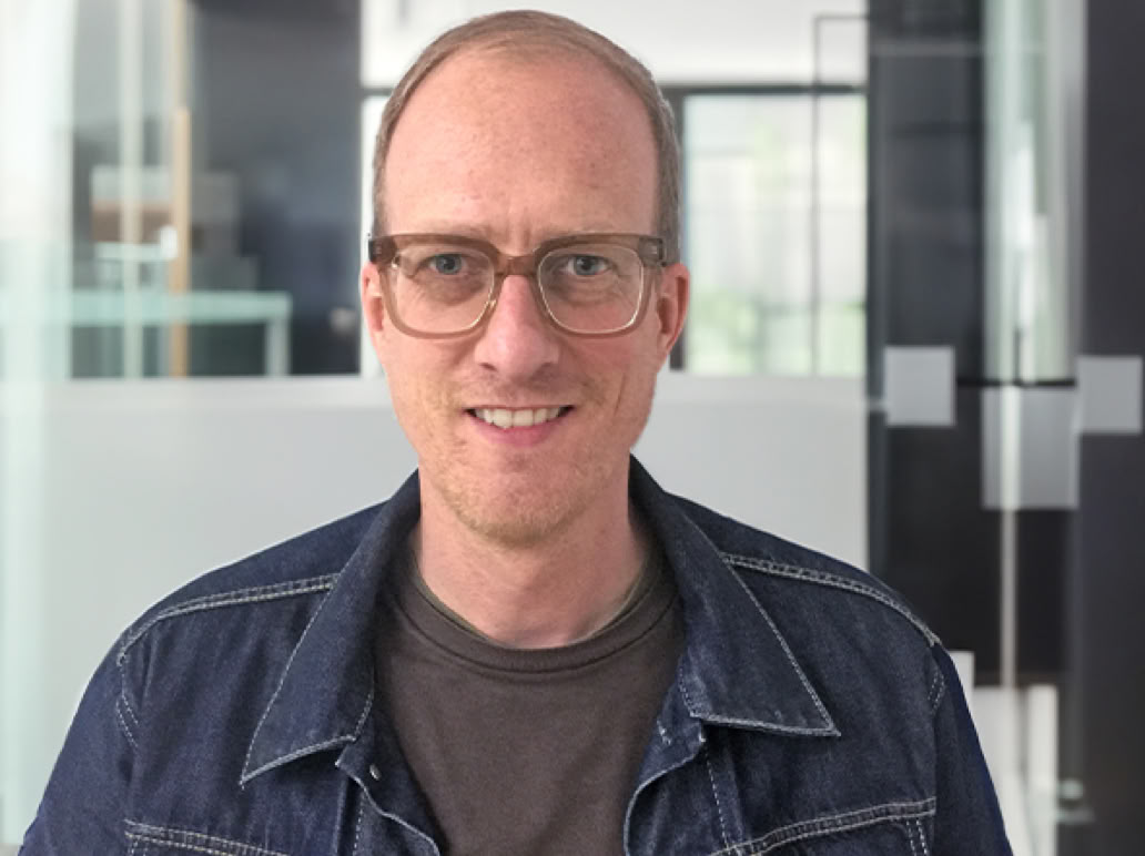 Ein Mann mit Brille und Jeansjacke steht in einem modernen Büro mit Glastüren im Hintergrund.
